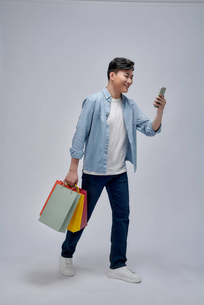 Giovane asiatico uomo con shopping bags sta usando un cellulare e sorridente mentre fa shopping - Foto, immagini
