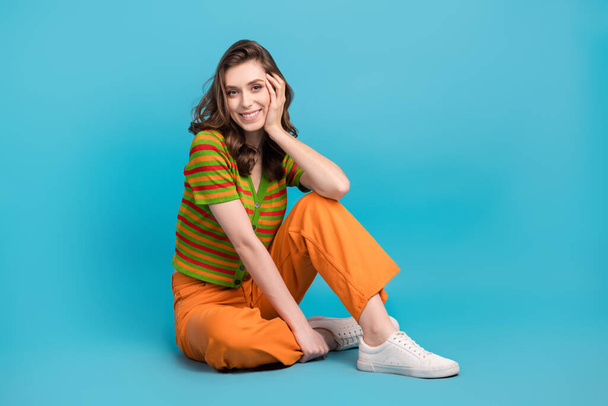 Full length photo of satisfied nice girl wear striped shirt trousers sitting hold arm on cheekbone isolated on blue color background. - Valokuva, kuva