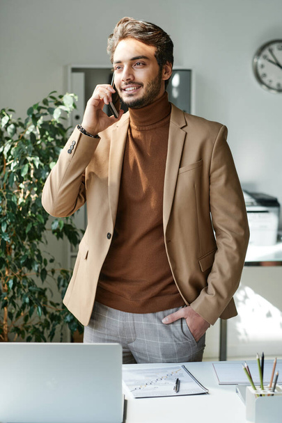 Smiling successful young Jewish businessman in stylish outfit standing at table and talking by mobile phone - Foto, immagini
