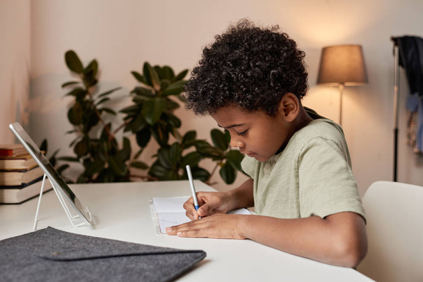 Serious curly-haired black boy sitting at desk and making notes in workbook during online lesson at home - Photo, image