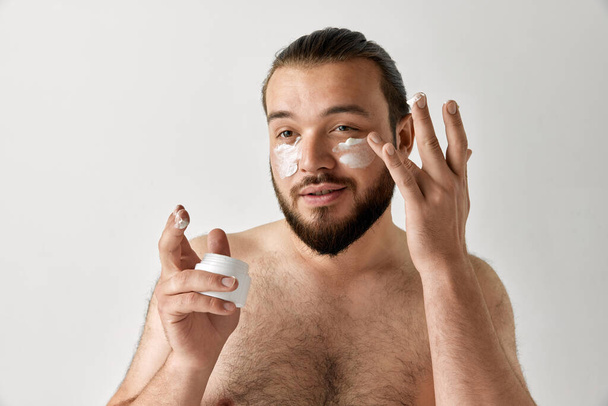 Retrato de hombre aplicando crema facial aislado sobre fondo gris estudio. Concepto de belleza, apariencia, cuidado corporal, salud y juventud. - Foto, imagen