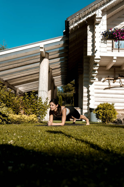 Eine durchtrainierte, hübsche Frau in Sportkleidung meditiert, während sie in einer Lotusposition auf dem grünen Gras in der Nähe ihres Hauses sitzt. junge Frau macht Yoga-Übungen zu Hause im Freien  - Foto, Bild