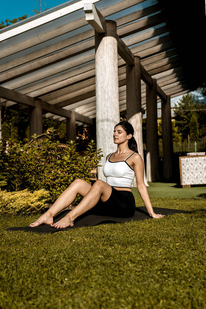 A fit, pretty woman in sports clothes meditates while sitting in a lotus position on the green grass near her house. young woman doing yoga exercises at home outdoors  - Foto, Imagem