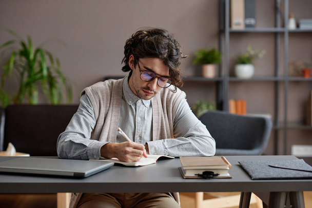 Horizontal medium portrait of young adult man wearing comfortable casual outfit and eyeglasses sitting at desk writing something in notebook - Foto, Bild