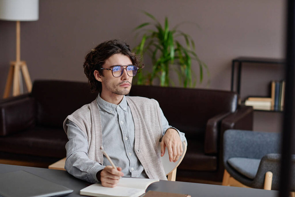 Horizontal medium portrait of handsome young adult psychotherapist wearing stylish casual outfit with eyeglasses sitting at desk looking away - Photo, image