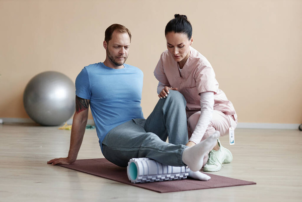 Пациент, сидящий на тренажерном коврике и тренирующийся во время реабилитации вместе с терапевтом - Фото, изображение