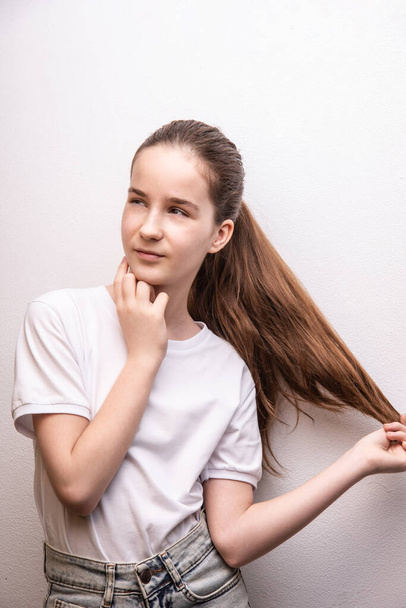 σκεπτικό κορίτσι αγγίζοντας παχιά υγιή μαλλιά, να κοιτάξουμε μακριά, φορώντας λευκό T-shirt πάνω από λευκό χρώμα υφή τοίχου - Φωτογραφία, εικόνα