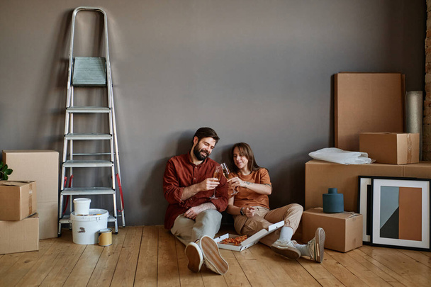 Horizontal shot of young man and woman sitting together on floor eating pizza and drinking champagne celebrating moving to loft apartment - Foto, Imagem