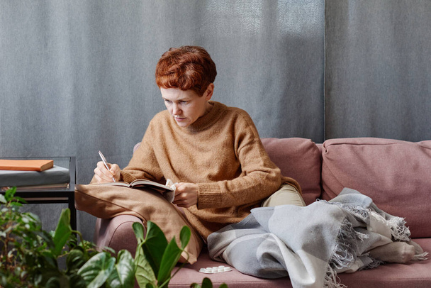 Horizontal shot of mature Caucasian woman having cold staying at home working on something making notes in notebook - Zdjęcie, obraz