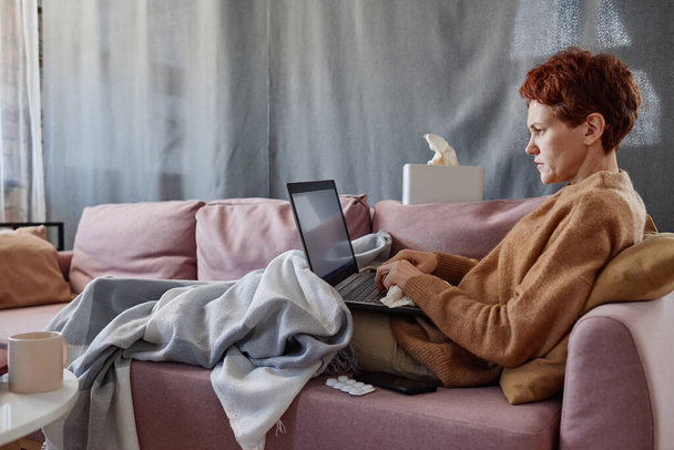 Horizontal shot of mature Caucasian woman suffering cold staying at home texting something on laptop - Fotografie, Obrázek
