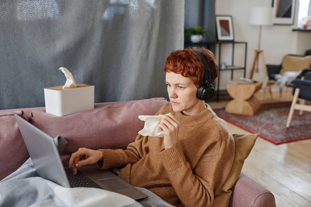 Horizontal shot of mature Caucasian woman having influenza staying at home sitting on sofa watching something on laptop - Photo, Image