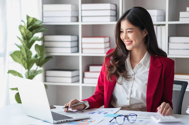 Азиатская бизнесвумен в красном костюме рассчитывает продажи компании с калькулятором, ноутбуком и планшетом на столе Интерьер офиса деловой женщины на современном рабочем месте. - Фото, изображение