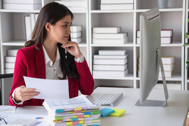 Азіатська бізнес-леді в червоному костюмі обчислює продажі компанії з калькулятором, ноутбуком і планшетом на столі Інтер'єр офісу бізнес-леді на сучасній робочій станції. - Фото, зображення