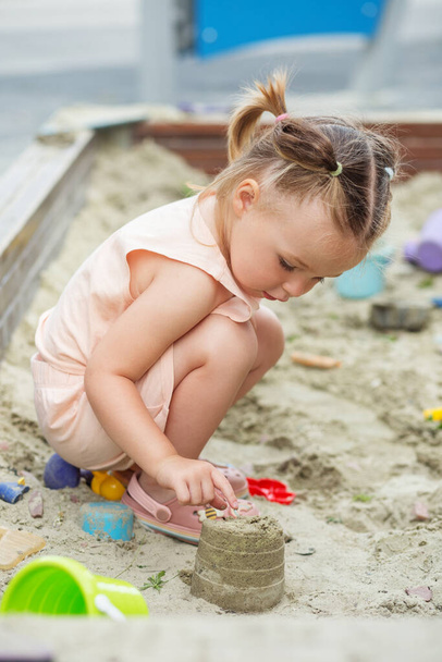 Pieni tyttö leikkii hiekkalaatikossa leikkikentällä ulkona. Pikkulapsi leikkii hiekkamuoteilla ja tekee mudpiiraita. Luova ulkoilu lapsille. - Valokuva, kuva