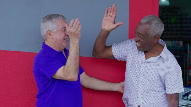 Zwei fröhliche ältere Freunde, die sich umarmen und feiern, während die High-Five draußen auf dem Bürgersteig stehen. Fröhliche afroamerikanische und kaukasische Kameradschaft authentische Interaktion - Foto, Bild