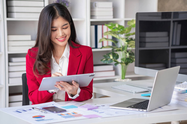 Азіатська бізнес-леді в червоному костюмі обчислює продажі компанії з калькулятором, ноутбуком і планшетом на столі Інтер'єр офісу бізнес-леді на сучасній робочій станції. - Фото, зображення