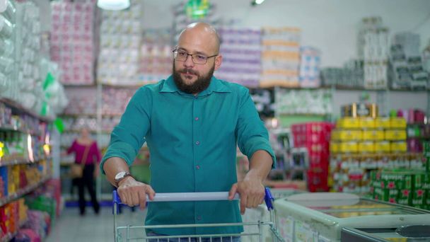Mužský zákazník Prohlížení produktů v obchodě s potravinami ulička, tlačení nákupní košík, zobrazování spotřebitelského životního stylu návyky - Fotografie, Obrázek