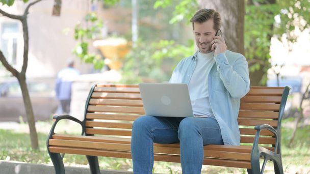 Молодой человек разговаривает по телефону во время использования ноутбука на открытом воздухе - Фото, изображение