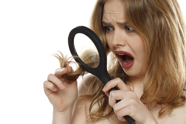 Ziemlich schockierte Frau mit Lupe, die ihr Haar auf weißem Studiohintergrund betrachtet - Foto, Bild