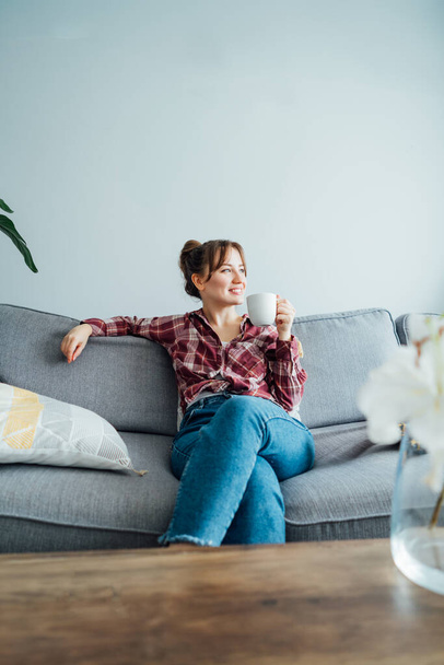 Young smiling woman sitting on sofa and looking side up while drinking coffee. Young brunette woman relaxing after housekeeping, home cleaning. Portrait of relaxed female resting at home. Vertical - Fotó, kép