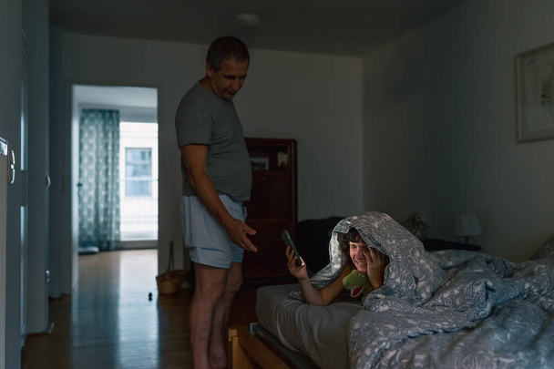 Táta a dcera sedí v pokoji pro teenagery v posteli s chytrými telefony, dcerou křičí, vyjadřuje nelibost, emocionálně zavírající oči, v noci. Generační konflikt, hádka mezi otcem a dcerou. - Fotografie, Obrázek