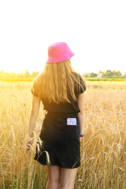Vista trasera de una chica bastante alta de pie en el campo de trigo amarillo. Día soleado. Sombrero panama rosa. Vertical. Teléfono púrpura en el bolsillo. - Foto, imagen