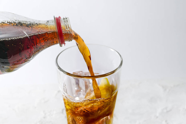 Pouring fresh cola from a bottle into a drinking glass with ice cubes and lemon slice, refreshing drink against a light gray background, copy space, selected focus, narrow depth of field - Photo, image