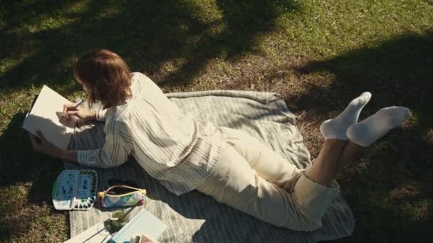 Μια ενήλικη, χαλαρή γυναίκα ξαπλωμένη στην κουβέρτα του πάρκου και ζωγραφίζοντας στο τετράδιο της. Μεσαίο πλάνο - Πλάνα, βίντεο