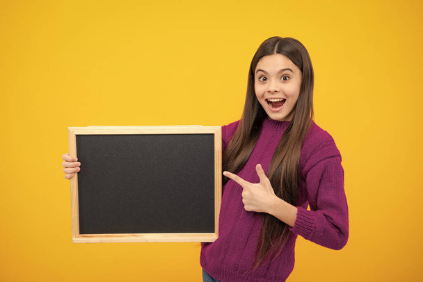 Teenager younf school girl holding school empty blackboard isolated on yellow background. Portrait of a teen female student. Copy space, mockup advertisement - Фото, изображение