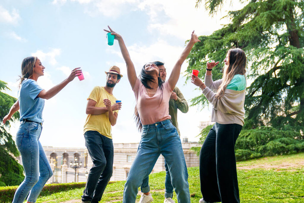 Levendig verjaardagsfeest in stadspark: 5 vrolijke, etnisch diverse vrienden dansen met kleurrijke feestbekers, creëren een levendige sfeer. - Foto, afbeelding