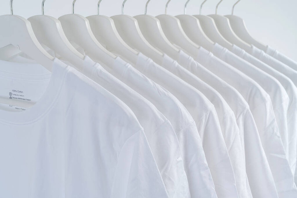 Крупный план белых футболок на вешалках, коллекция белых футболок висит на деревянной вешалке, на фоне одежды - Фото, изображение