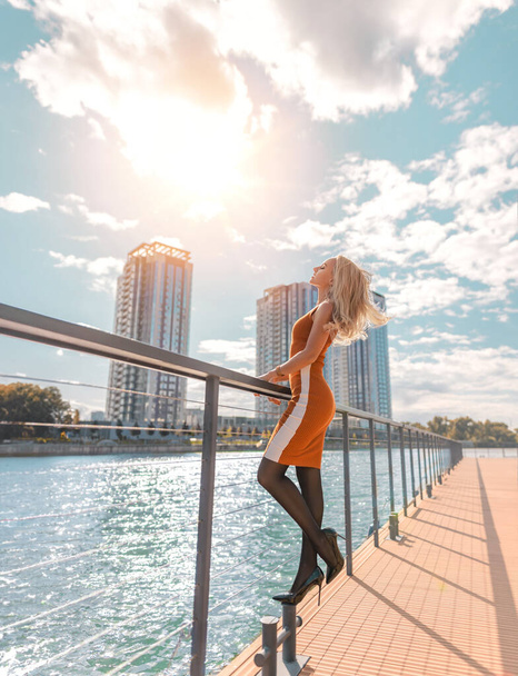 Beautiful blonde girl in orange dress with perfect legs in pantyhose and shoes with high heels posing outdoor on the bridge with the Sun. - Foto, imagen