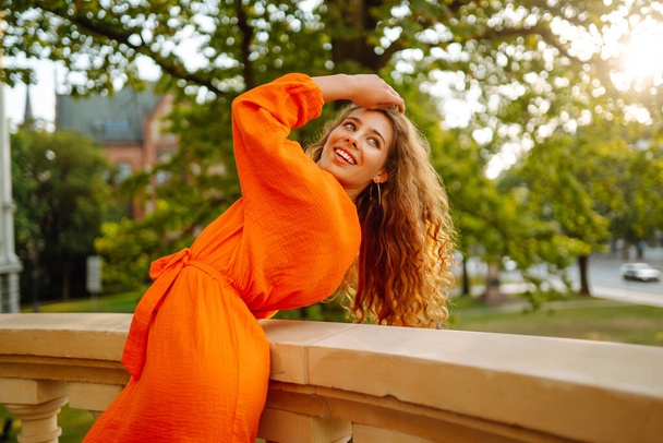 Happy young model in a bright dress poses against the backdrop of a city landscape. Curly woman enjoys the beautiful sunny weather of the city. Lifestyle concept. Photo on the street. - Фото, зображення