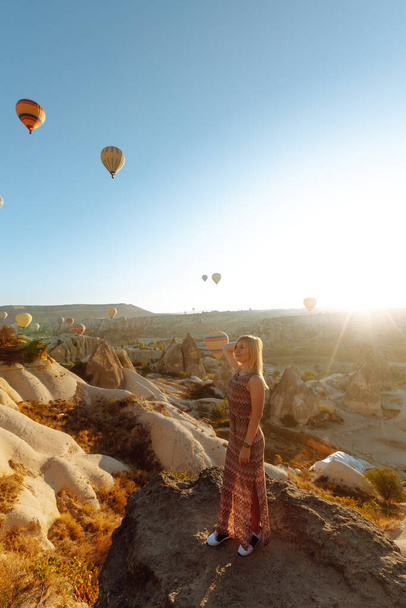 Attractive young woman in hat stands on mountain with her back, enjoying view of flying hot air balloons. Enjoy the moment. Concept of freedom, active lifestyle. Famous tourist region Cappadocia. - Photo, Image