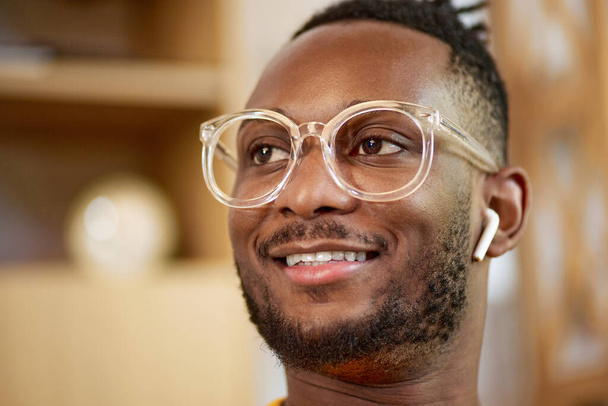 Close-up portrait of young african man in earphones and glasses, smiling. Blurred home interior on background. Student, worker. Concept of business and education, freelance job, modern lifestyle - Foto, Bild