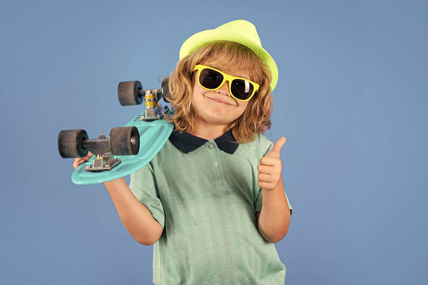 Studio portrait of stylish cute young child boy with skateboard isolated against blue background. Kid skater. Fashion kid. Stylish little child wearing a summer clothes, sunglasses and hat - Fotografie, Obrázek
