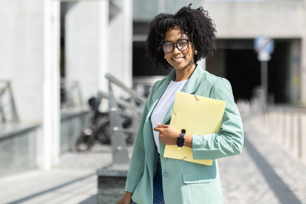 Retrato de una alegre y atractiva joven afroamericana en gafas casuales inteligentes con bloc de notas en la mano empresaria gerente posando al aire libre junto al edificio de oficinas, espacio para copiar - Foto, Imagen