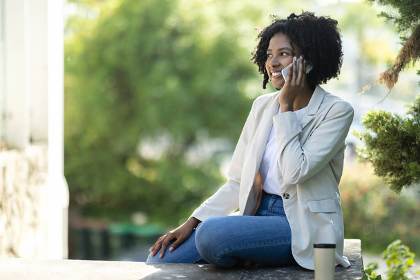 Приваблива усміхнена молода афроамериканська корпоративна бізнес-леді в розумному повсякденному вбранні має телефонний дзвінок, сидить на сходах у парку на вулиці, п'є каву, копіює простір - Фото, зображення