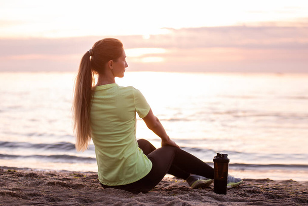 Young woman in sportswear is sitting on the seashore at sunrise. Girl is resting after workout, replenishes her water balance, drinks from a sports bottle, looks at the water. Morning outdoor fitness classes. - Foto, Imagen
