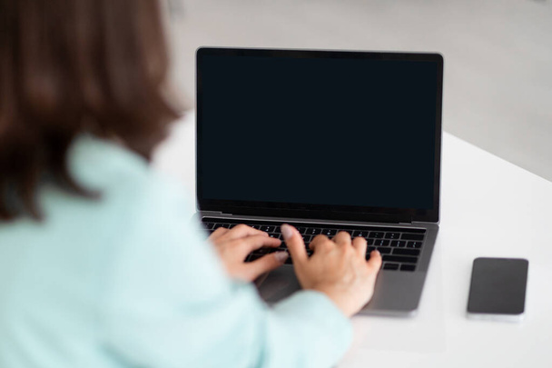 Millennial kaukasische Dame tippt am Computer mit leerem Bildschirm am Arbeitsplatz mit Smartphone im Bürointerieur, abgeschnitten. Geschäft mit Gerät, Managerarbeit, Studienapp und Website - Foto, Bild