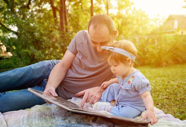 Widok z bliska ojca czytającego książkę z dzieckiem na podwórku. Piękne rodzinne przytula i całuje dziecko wspierane przez kochających rodziców. Przycięty portret uroczej dziewczynki w sukience. - Zdjęcie, obraz