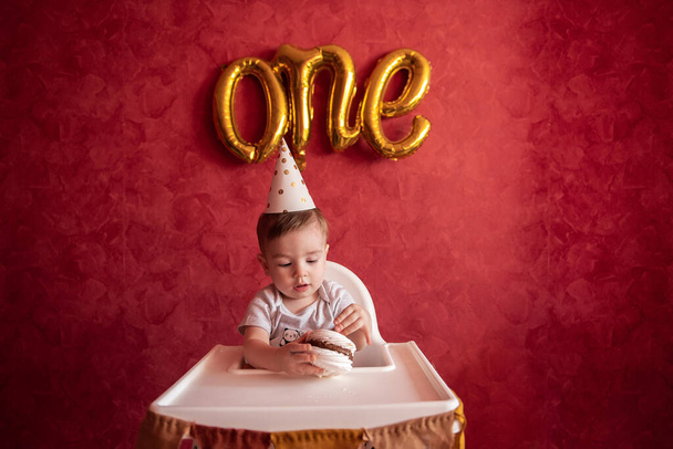 Little boy of one year old in party hat sits in childrens chair, eating birthday cake, Meringue, blows out candle. Kid on red isolated background, on textured wall foil gold balloons One. Home party - Фото, изображение