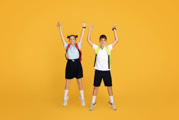 Cheerful caucasian teenager girl and boy pupils with backpacks jumping, freeze in air, celebrating victory, have fun, isolated on orange studio background. Study, school education, ad and offer - Foto, imagen