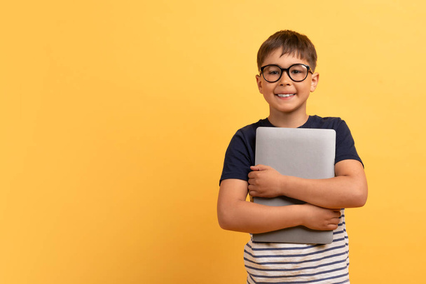 Cheerful handsome cute guy school aged kid boy in casual t-shirt and eyeglasses embracing computer laptop and smiling, isolated on yellow background, copy space. Gadget addiction in children - Photo, Image