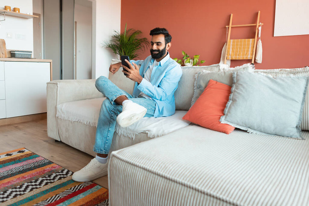 Lächelnder Inder, der die Online-Freizeit mit dem Handy genießt, zu Hause SMS schreibt und kommuniziert, auf dem Sofa sitzt und sich am Wochenende entspannt. Leerer Raum für Text. Gadgets und Internet - Foto, Bild