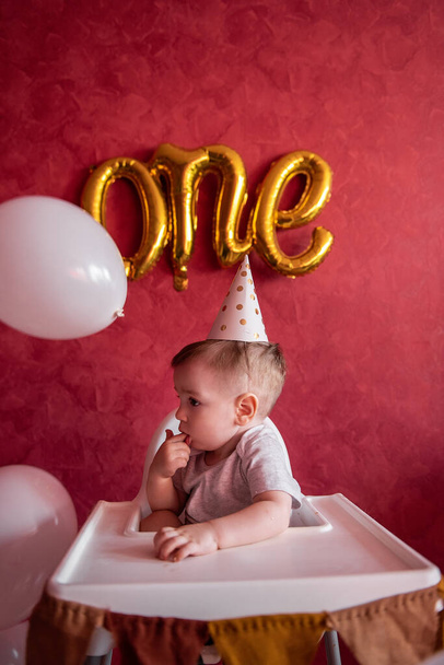 Portrait of Little boy of one year in festive hat sits in childrens chair with fabric flags. Kid on red isolated background with foil gold balloons One, on textured wall celebrating holiday at home - Φωτογραφία, εικόνα