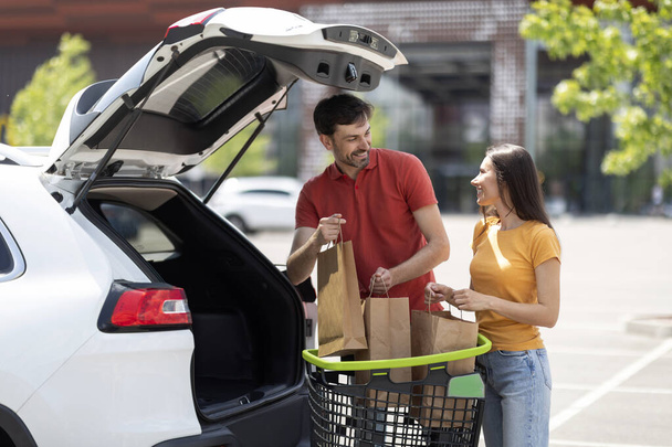 Супруги тысячелетия упаковывают багажник машины с продуктами. Веселый любящий молодой человек и женщина в случайном стоянии на авто с корзиной полный покупок, возвращаясь домой из супермаркета - Фото, изображение