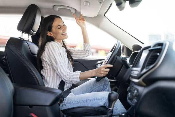 Happy cheerful pretty long-haired young woman wearing casual outfit driver sitting in front seat, holding auto steering wheel, driving her brand new nice car, listening to music, have fun, copy space - Photo, Image