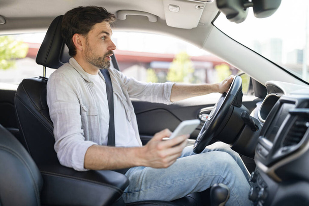 Distracted driving concept. Shocked scared handsome young man in casual outfit driver using smartphone while driving auto, got car accident. Guy holding phone in his hand, crushed automobile - Photo, Image