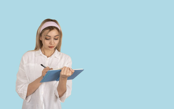 Taking notes, portrait of teenage blonde caucasian girl taking notes. Making checklist. Young smart university student studying, writing on notebook. Isolated blue background copy space. - Photo, Image